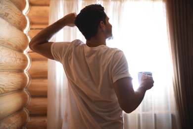 Photo of Man with drink stretching near window indoors. Lazy morning