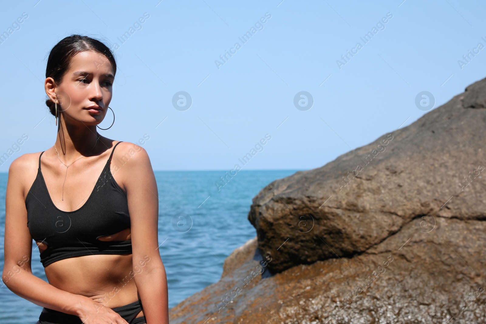 Photo of Beautiful young woman in stylish bikini sitting on rock near sea, space for text