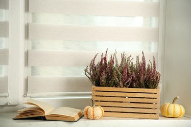 Photo of Beautiful heather flowers in crate, book and pumpkins on white windowsill indoors