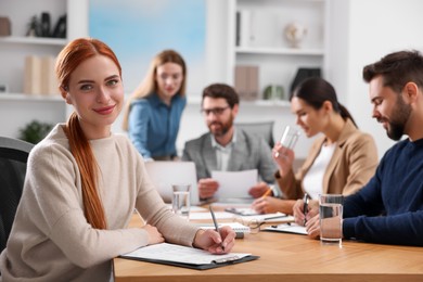 Photo of Team of employees working together in office. Happy woman with pen and clipboard at table indoors