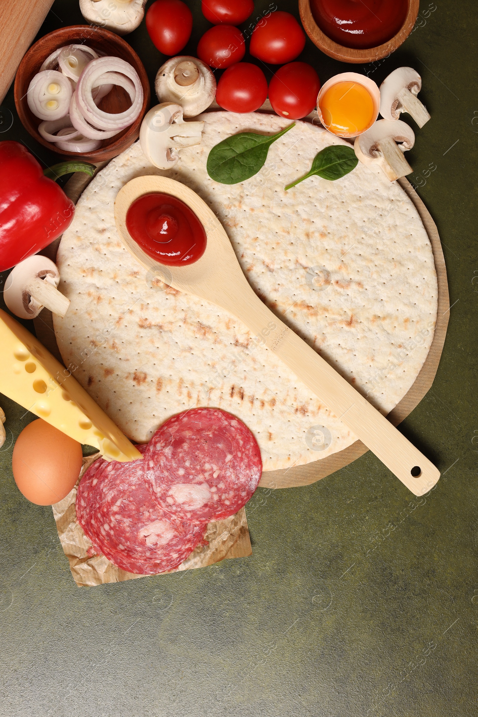 Photo of Pizza base, spoon with tomato sauce and products on green textured table, flat lay