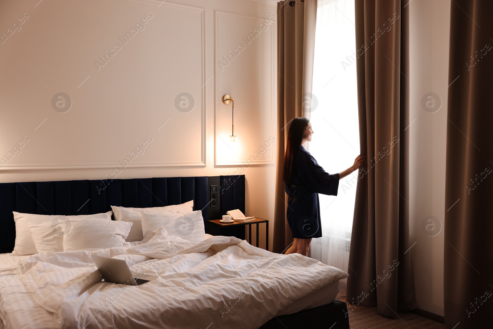 Photo of Beautiful young woman wearing silk robe near window in hotel room