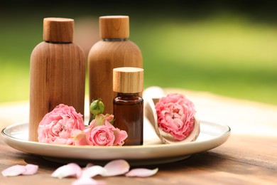 Bottles of rose essential oil and flowers on wooden table outdoors, space for text