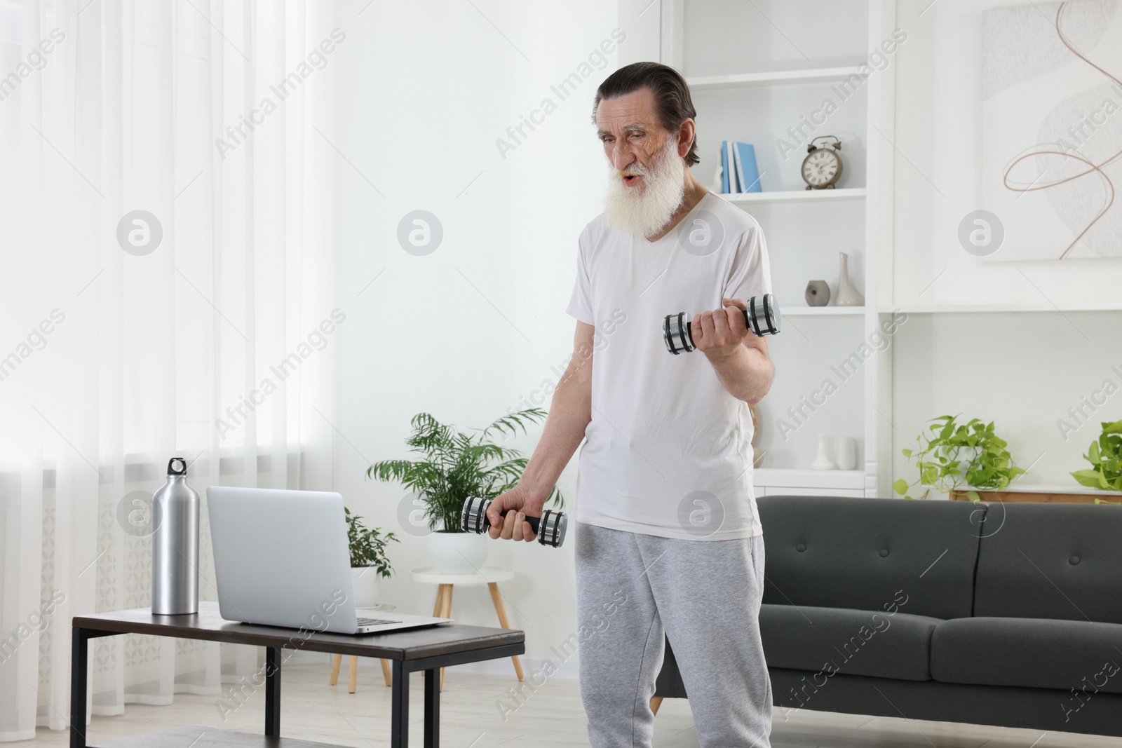 Photo of Senior man exercising with dumbbells while watching online tutorial at home. Sports equipment