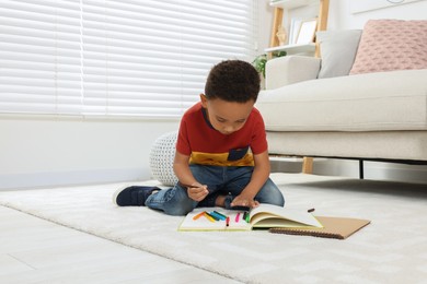 Cute African-American boy drawing in sketchbook with colorful markers on floor at home