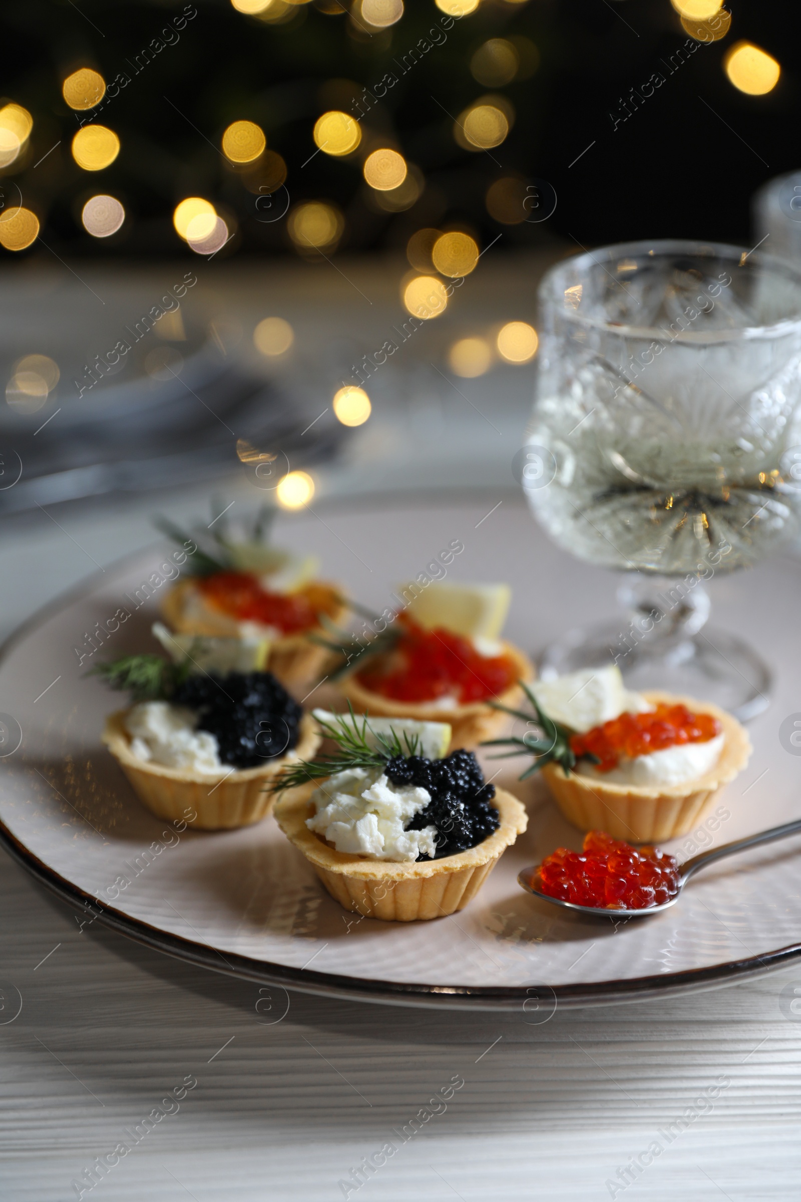 Photo of Delicious tartlets with red and black caviar served on white wooden table against blurred festive lights, closeup. Space for text