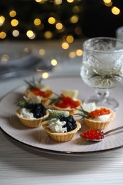 Delicious tartlets with red and black caviar served on white wooden table against blurred festive lights, closeup. Space for text