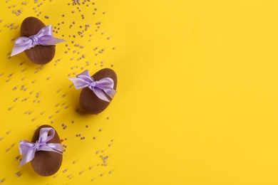 Photo of Sweet chocolate eggs with bows and sprinkles on yellow background, flat lay. Space for text