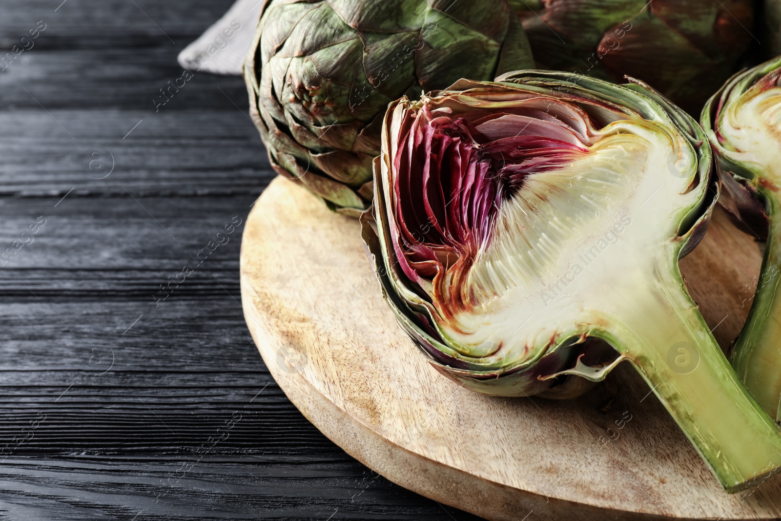 Photo of Cut and whole fresh raw artichokes on black wooden table, closeup. Space for text