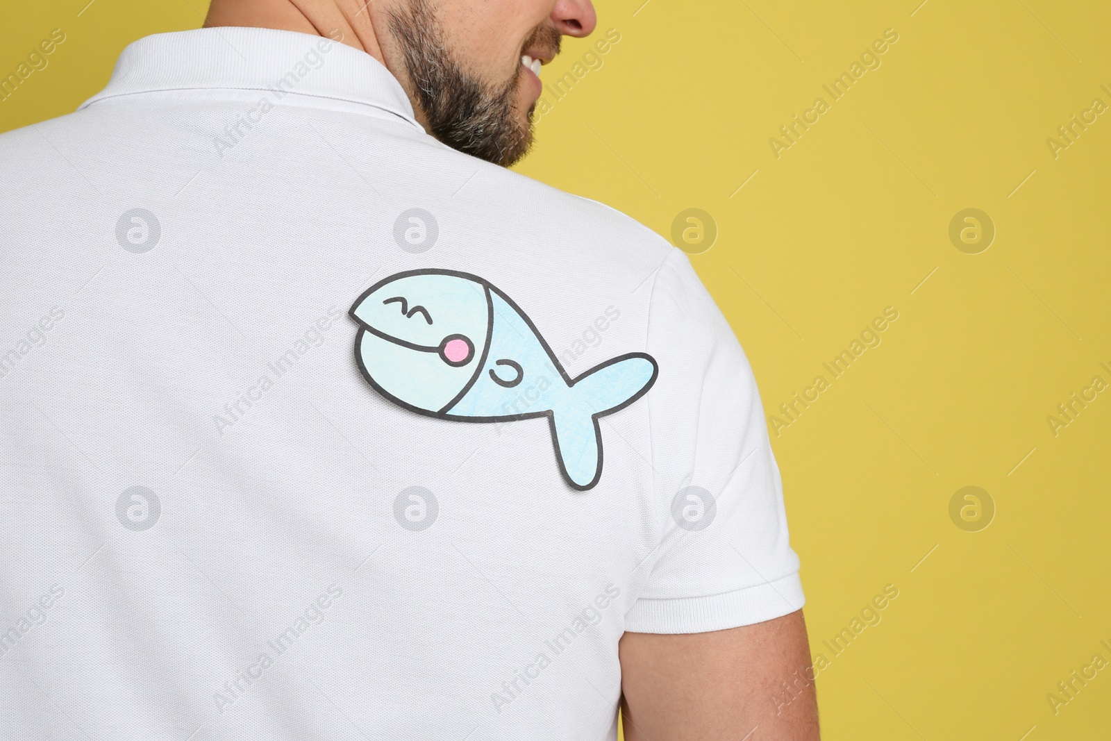 Photo of Man with paper fish on back against yellow background, closeup. April fool's day