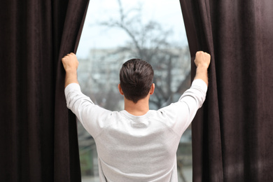 Man opening window curtains at home, back view