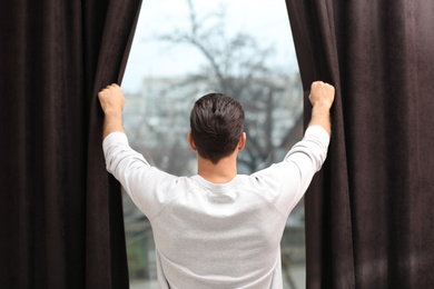 Photo of Man opening window curtains at home, back view