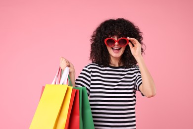 Happy young woman with shopping bags and stylish sunglasses on pink background