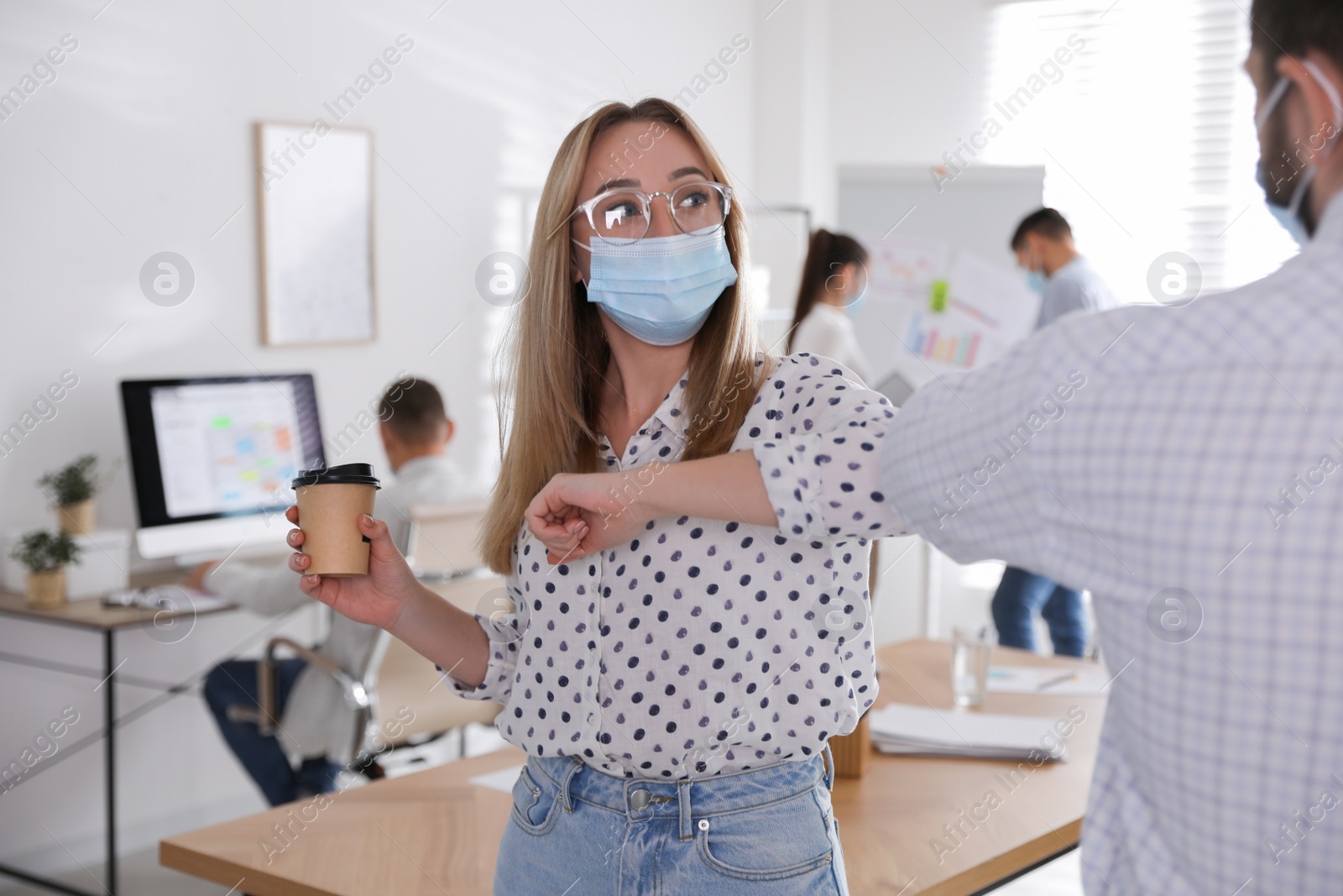 Photo of Coworkers with protective masks making elbow bump in office. Informal greeting during COVID-19 pandemic