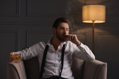 Photo of Handsome man with glass of whiskey smoking cigar in armchair at home