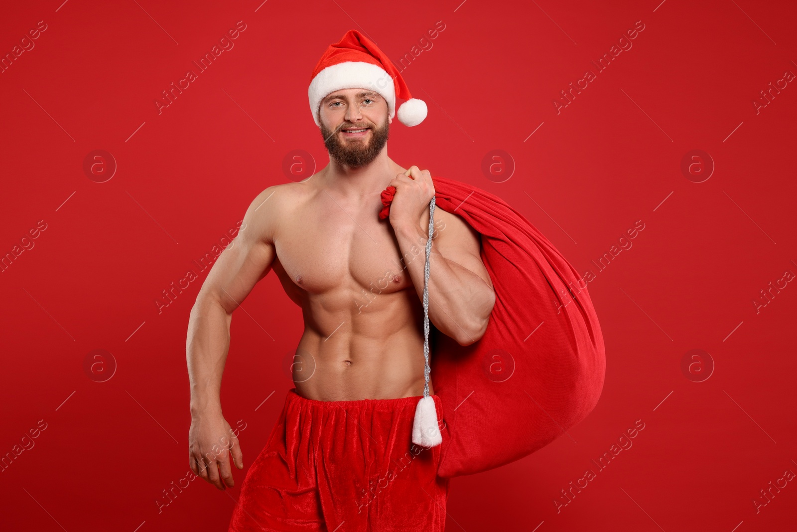 Photo of Muscular young man in Santa hat holding bag with presents on red background