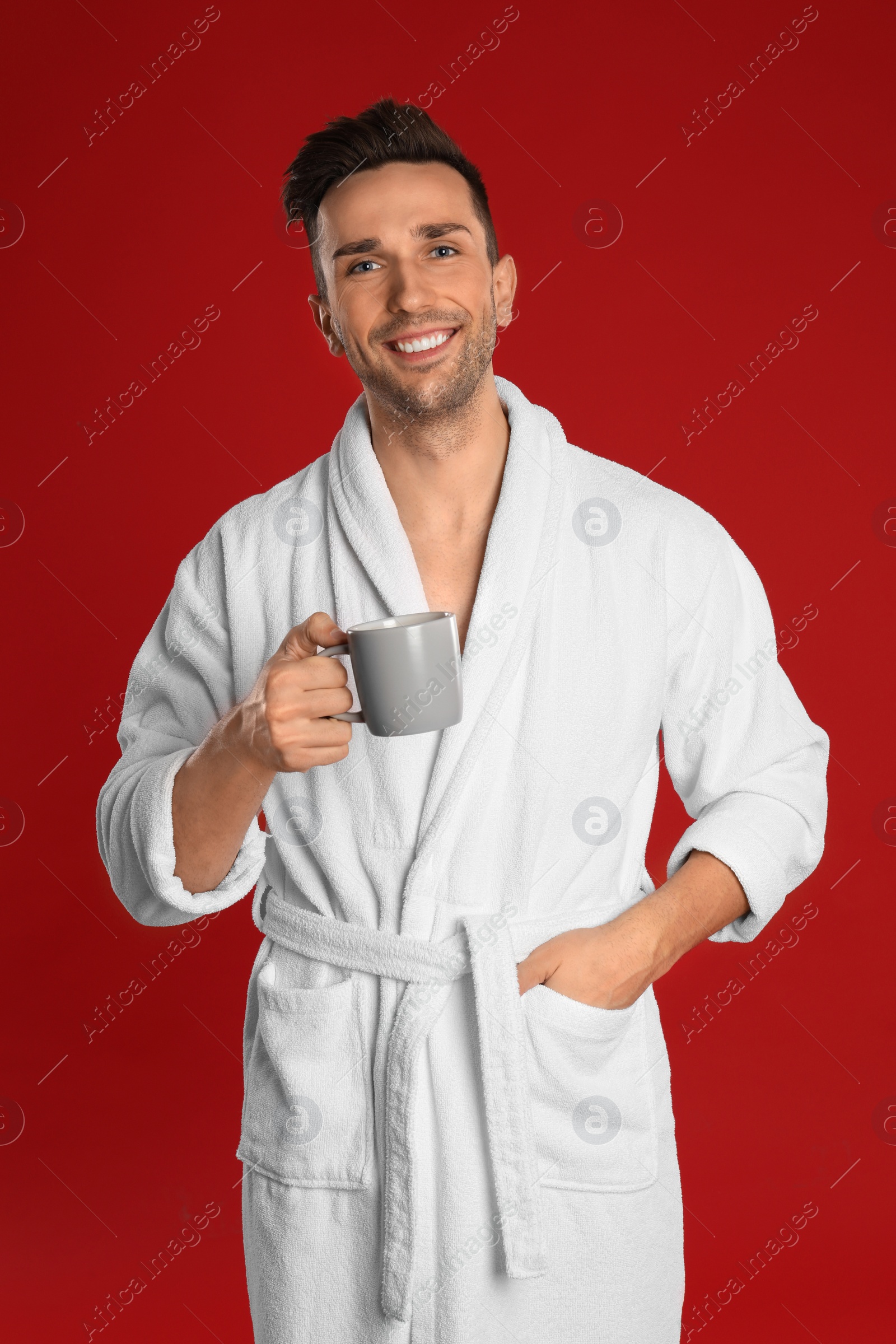 Photo of Happy young man in bathrobe with cup of coffee on red background