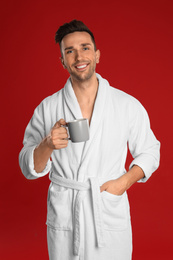 Happy young man in bathrobe with cup of coffee on red background