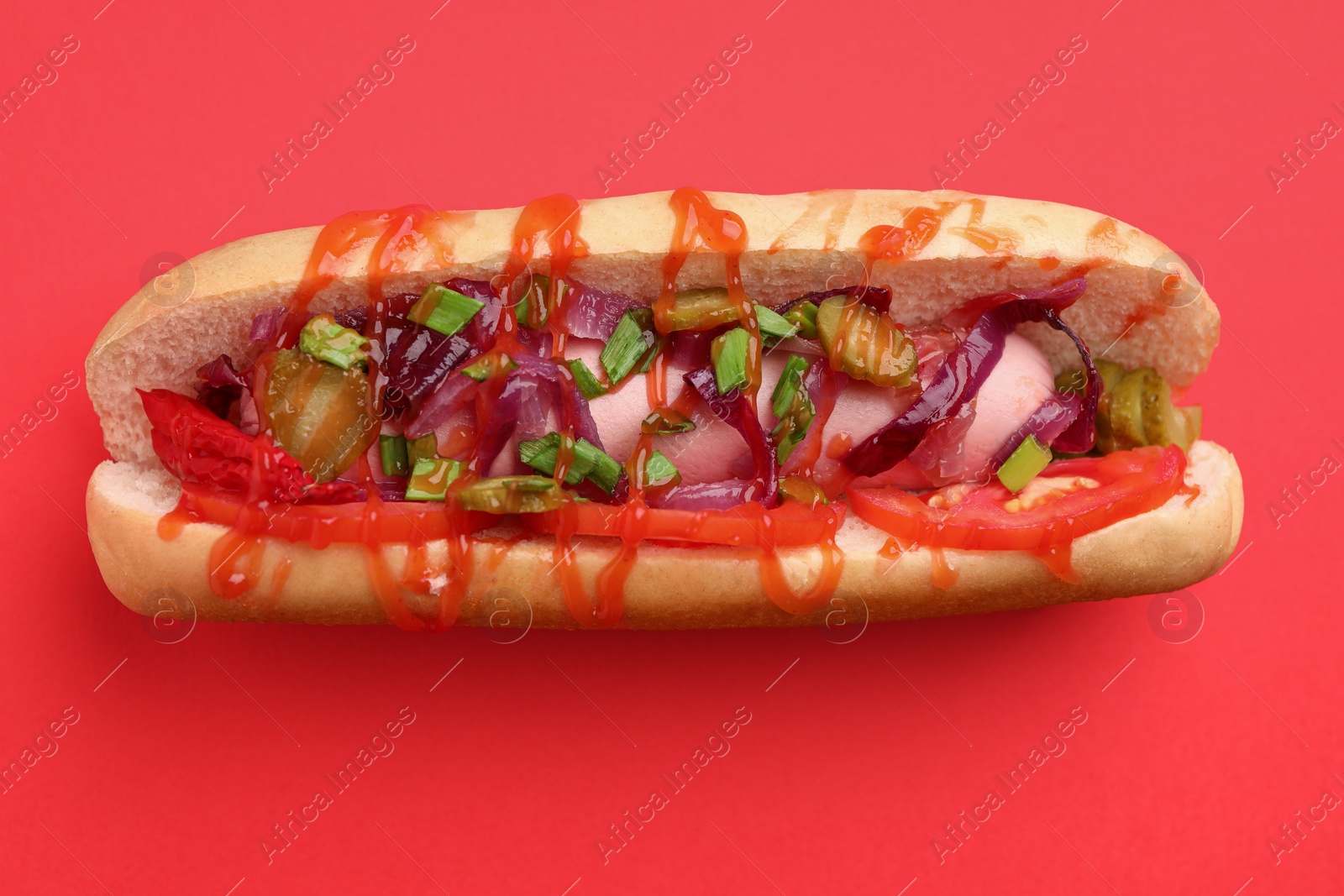 Photo of Tasty hot dog with onion, tomato, pickles and sauce on red background, top view