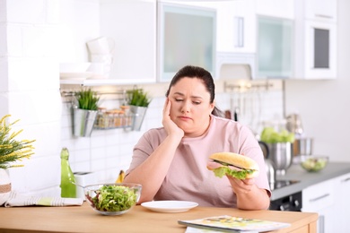 Photo of Sad overweight woman holding sandwich at table in kitchen. Healthy diet