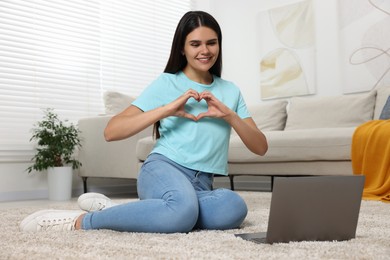 Photo of Happy young woman having video chat via laptop and making heart on floor in living room