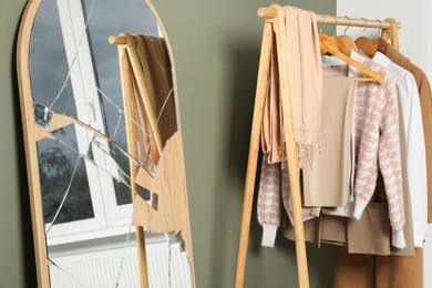 Broken mirror and wooden rack with clothes near olive wall indoors