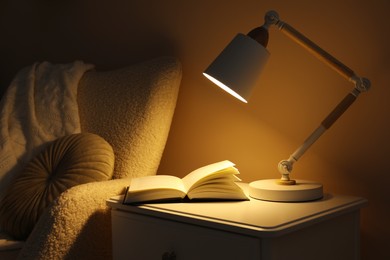 Photo of Stylish modern desk lamp and open book on white cabinet near armchair in dark room