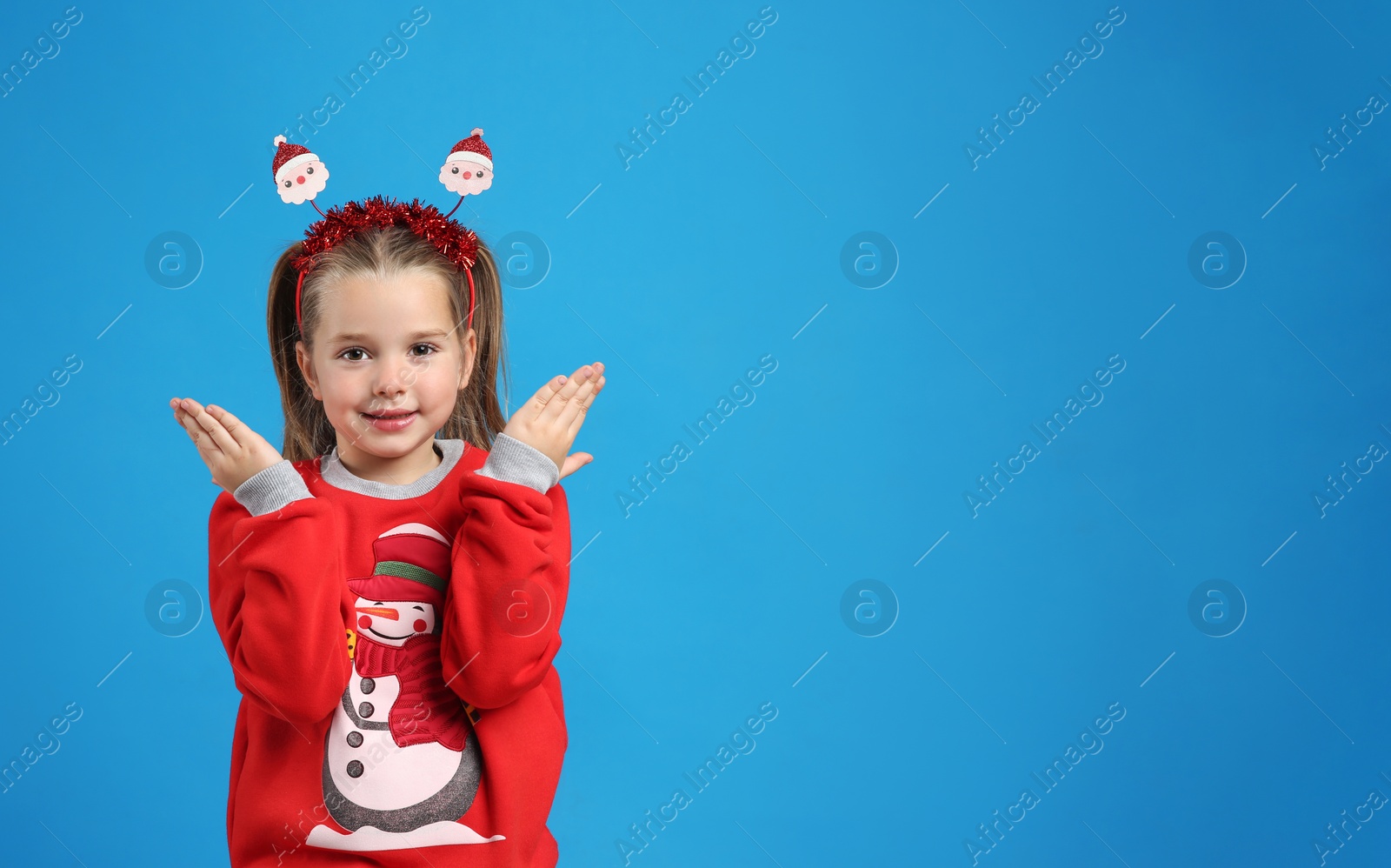 Photo of Cute little girl in Christmas sweater and Santa headband on blue background, space for text