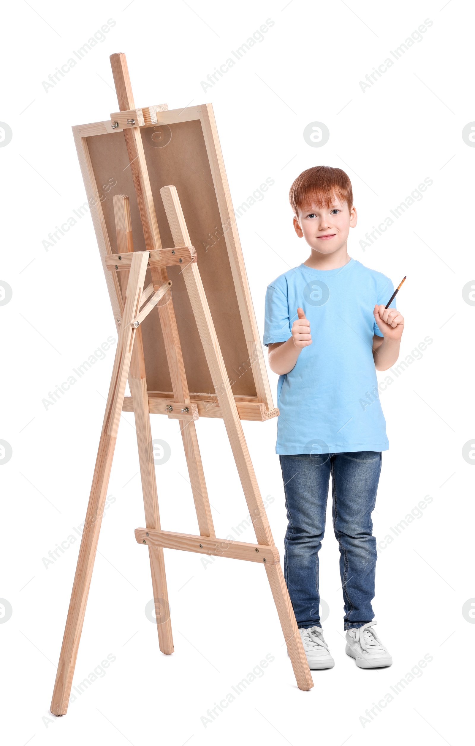 Photo of Little boy with brush near easel with canvas showing thumbs up against white background. Creative hobby