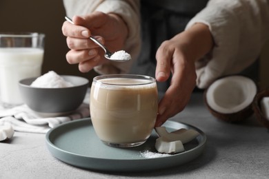 Woman putting coconut flakes into glass of coffee with milk at light grey table, closeup