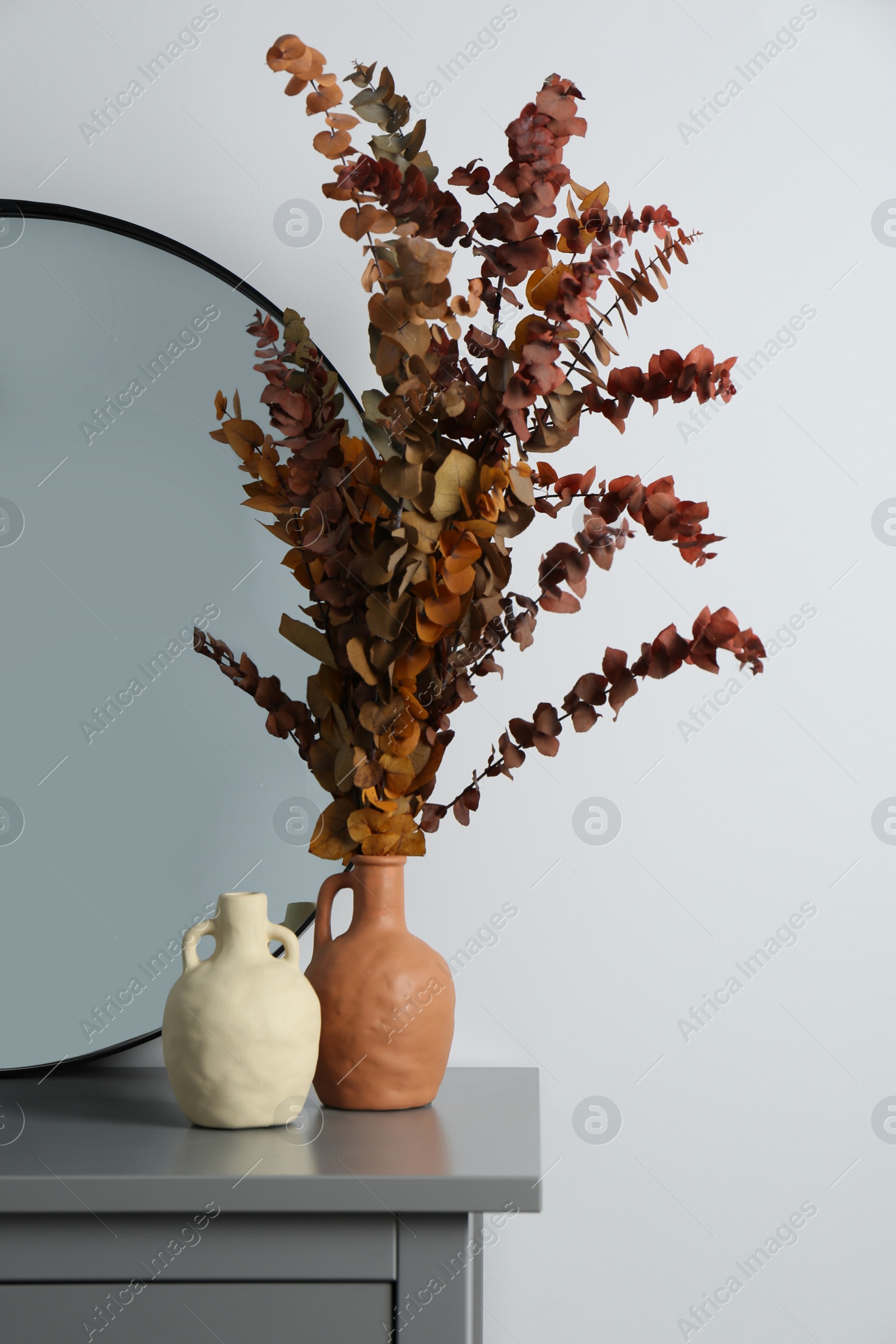 Photo of Vases, dried eucalyptus branches and stylish round mirror on grey table near white wall indoors. Interior design