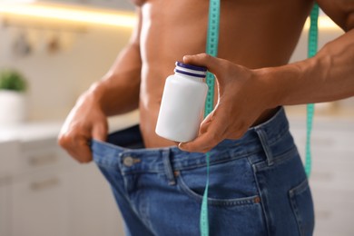 Athletic man with measuring tape and bottle of supplements wearing big jeans indoors, closeup. Weight loss