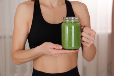 Photo of Young woman in sportswear with mason jar of fresh smoothie at home, closeup
