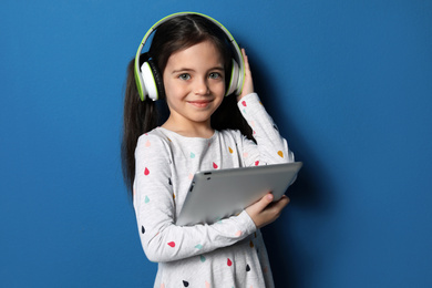 Photo of Cute little girl with headphones and tablet listening to audiobook on blue background