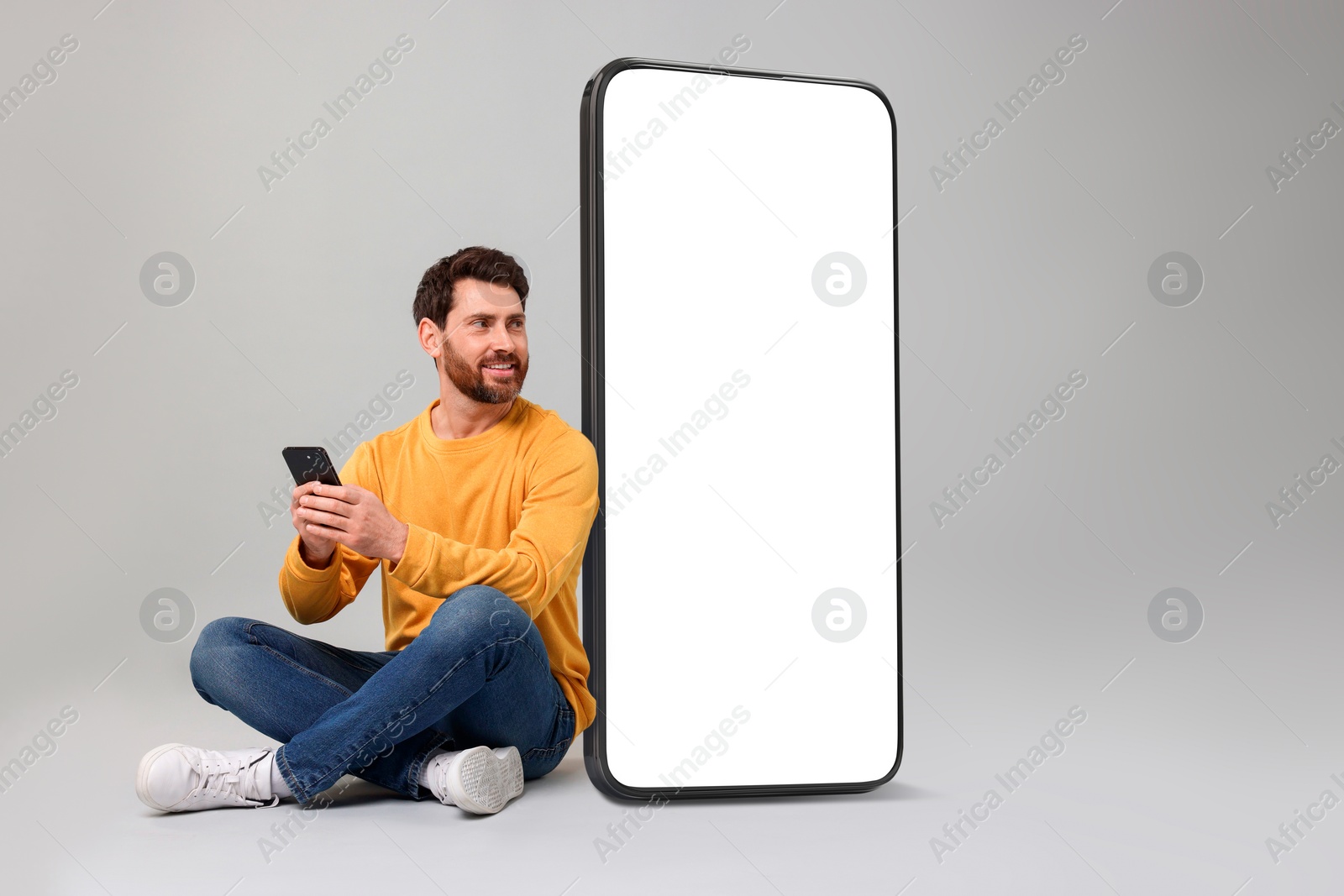 Image of Man with mobile phone sitting near huge device with empty screen on grey background. Mockup for design