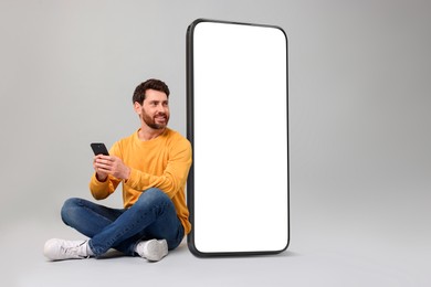 Man with mobile phone sitting near huge device with empty screen on grey background. Mockup for design