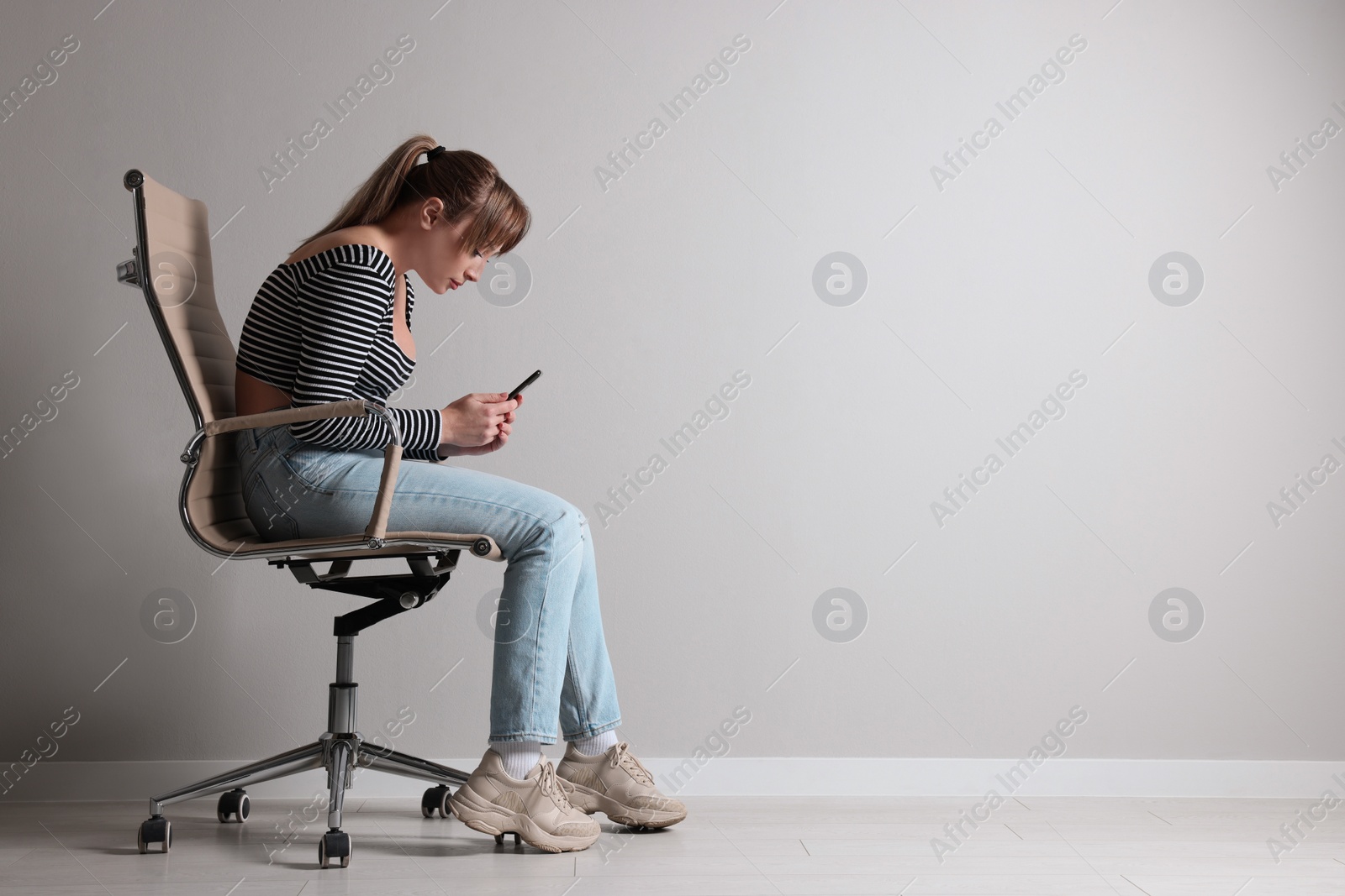 Photo of Young woman with poor posture using smartphone while sitting on chair near grey wall, space for text