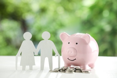 Pension savings. Figure of senior couple, piggy bank and coins on white table against blurred green background