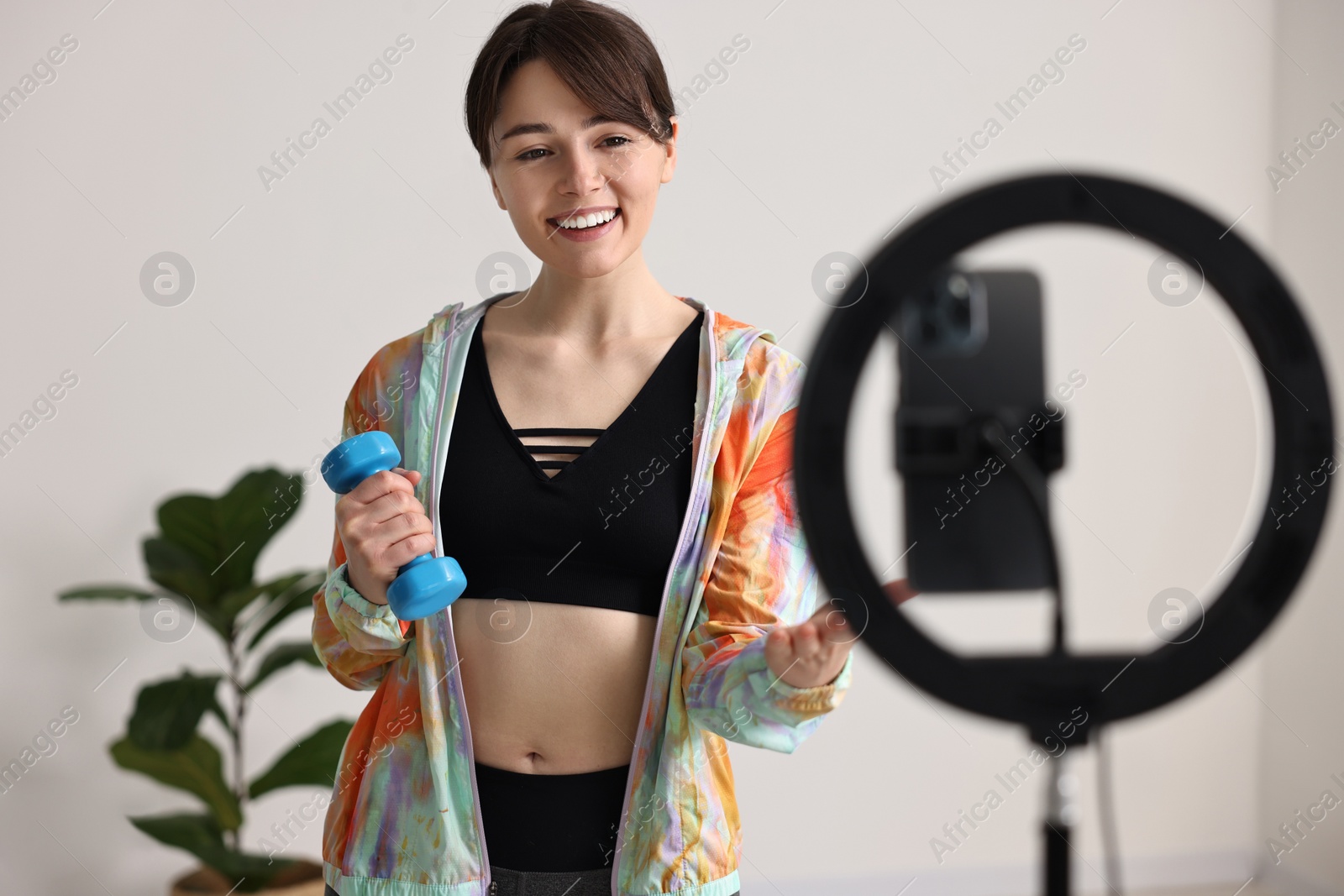 Photo of Happy sports blogger holding dumbbell while streaming online fitness lesson with smartphone at home