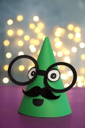 Photo of Funny handmade party hat with glasses and moustache on purple table against blurred lights, closeup