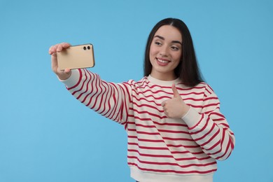 Photo of Smiling young woman taking selfie with smartphone and showing thumbs up on light blue background