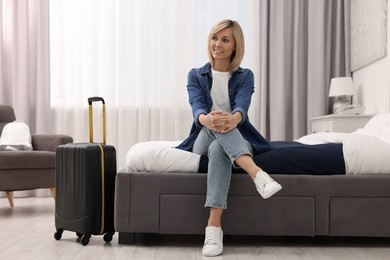 Smiling guest with suitcase relaxing on bed in stylish hotel room