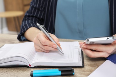 Photo of Woman taking notes while using smartphone at wooden table, closeup