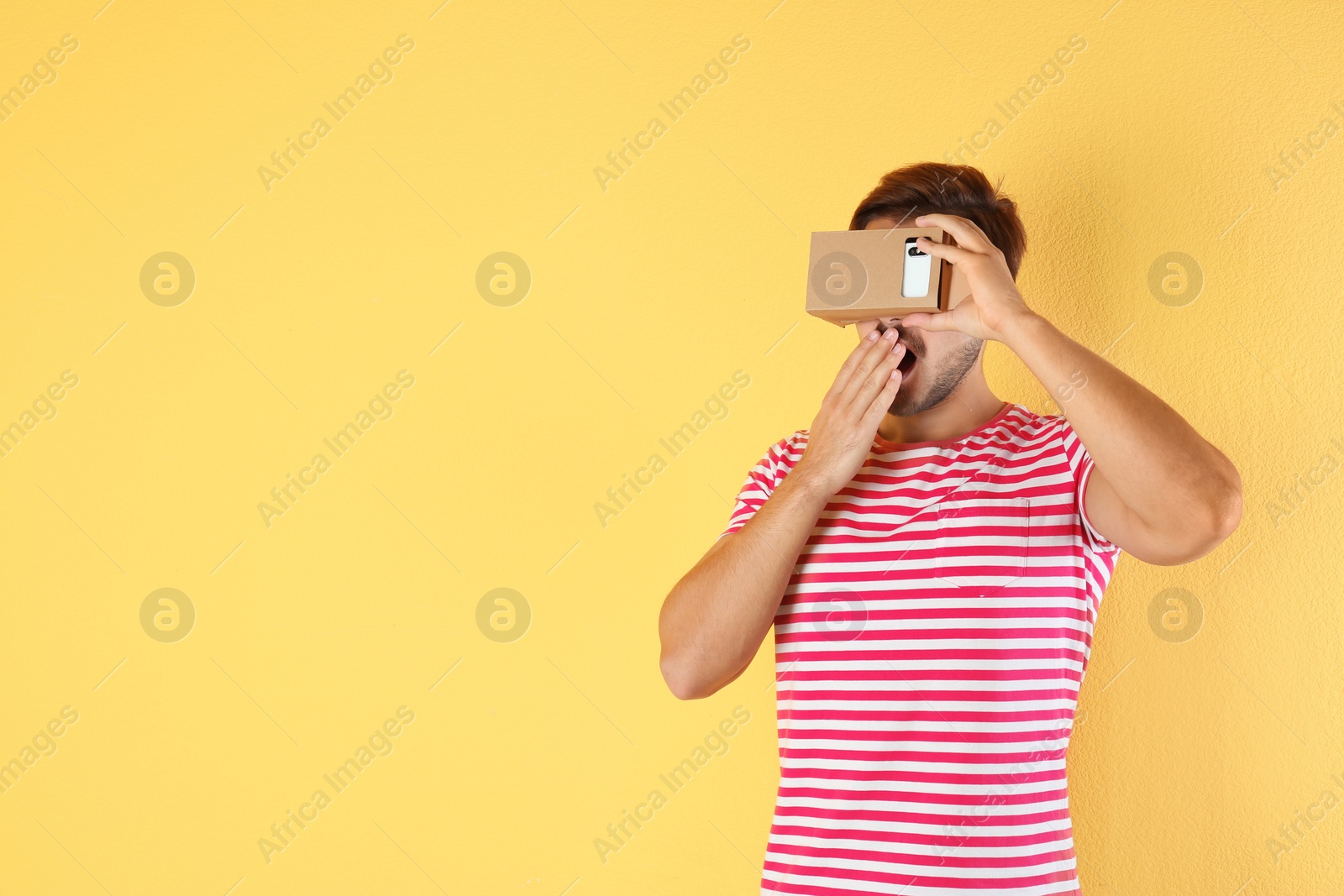 Photo of Young man using cardboard virtual reality headset on color background. Space for text