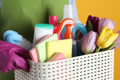 Photo of Spring cleaning. Woman holding basket with detergents, flowers and tools on orange background, closeup