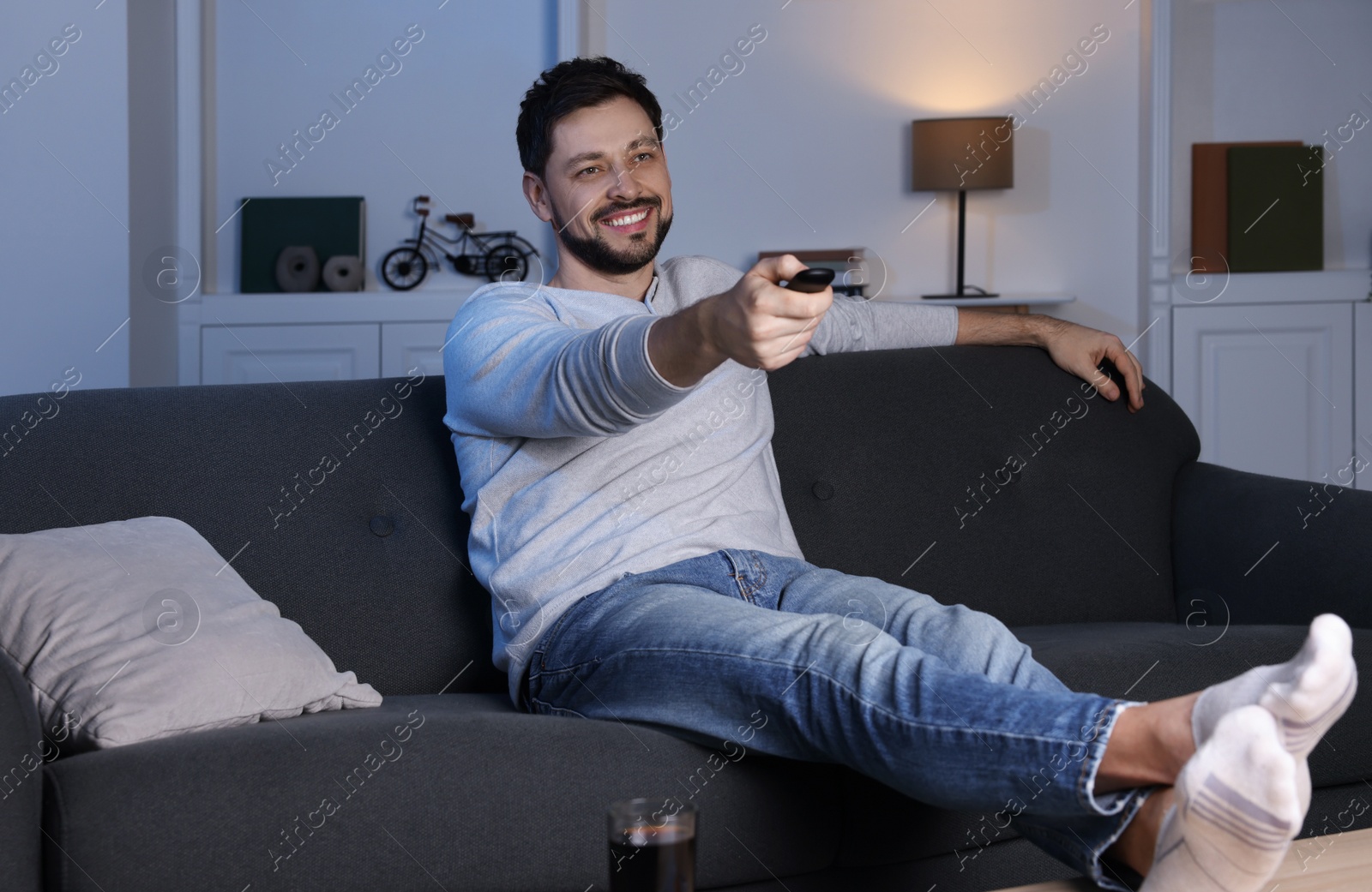 Photo of Happy man changing TV channels with remote control on sofa at home