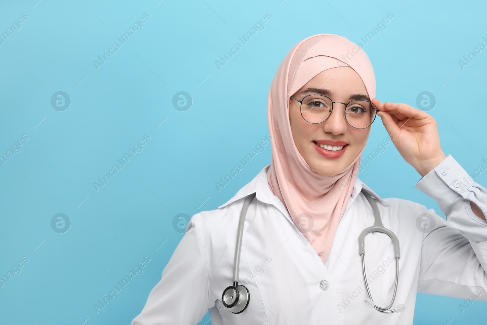 Photo of Muslim woman wearing hijab and medical uniform with stethoscope on light blue background, space for text