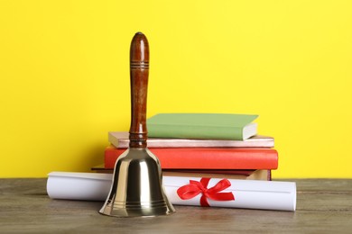 Photo of Golden school bell, diploma and books on wooden table against yellow background