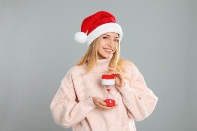 Photo of Woman in Santa hat with hourglass on grey background. Christmas countdown