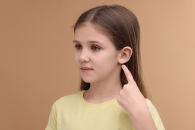 Hearing problem. Little girl pointing at ear on pale brown background